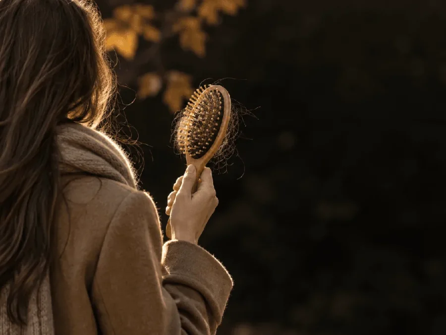 Autumn hair loss: woman in a camel coat holding a wooden brush with shed hairs in the bristles