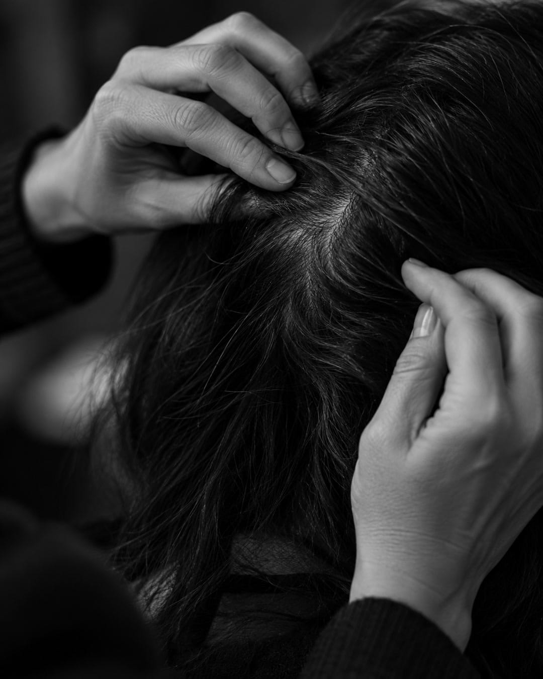 Black-and-white editorial scene of a professional's hands gently parting a client's dark hair to look at the scalp